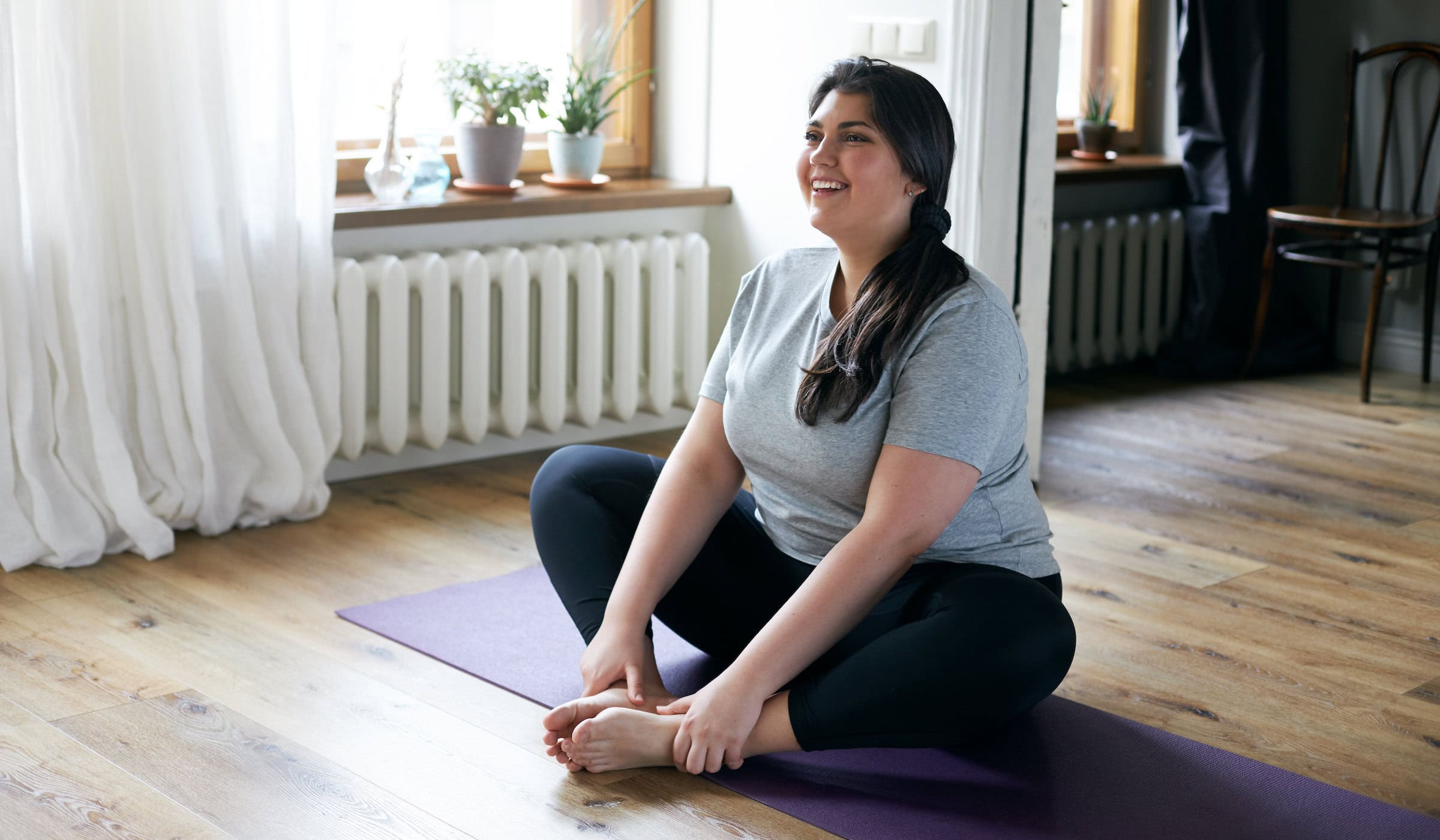 Cheerful attractive young overweight woman in activewear choosing healthy lifestyle, sitting on mat with hands on bare feet, doing butterfly yoga exercise, stretching thighs. Body shape and activity